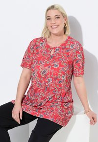 Červená tunika s krátkým rukávem a paisley vzorem, s výstřihem ve tvaru klíčky a volným střihem, v kombinaci s černými kalhotami. Hladká tkanina s barevnými akcenty.