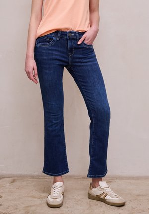 Frau trägt dunkelblaue Bootcut-Jeans, pfirsichfarbenes Top leicht eingesteckt und weiße Sneakers, stehend auf beigem Boden vor einer schlichten Wand.