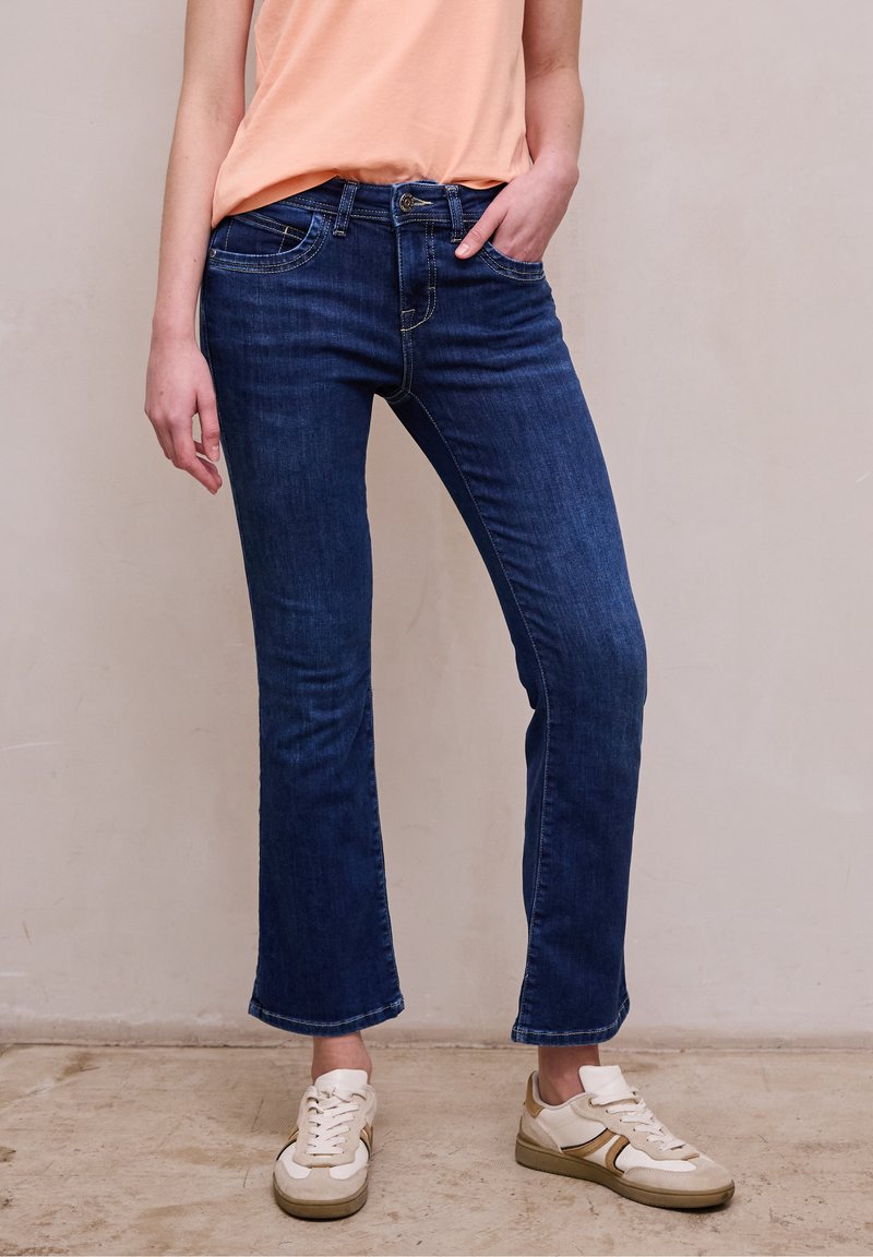 Kobieta ubrana w ciemnoniebieskie jeansy bootcut, brzoskwiniową koszulkę lekko wpuszczoną do spodni i białe trampki, stoi na beżowej podłodze na tle gładkiej ściany.