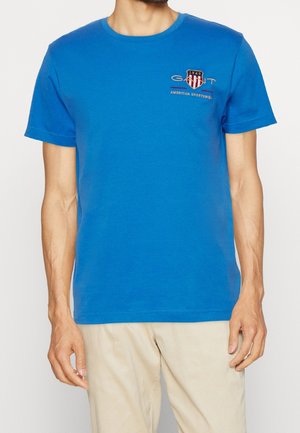Homme portant un t-shirt bleu à manches courtes avec un petit logo "GANT 1949 American Sportswear" sur la poitrine gauche et un pantalon beige.