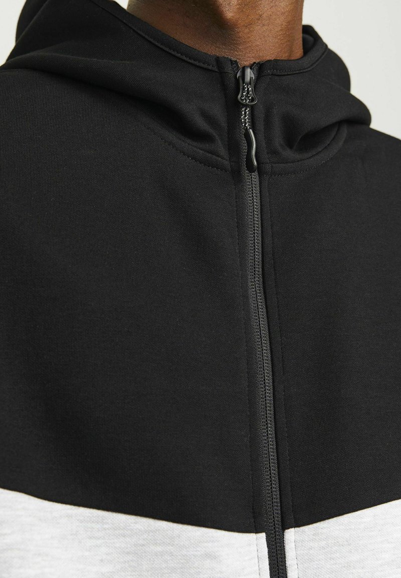 Felpa nera con cappuccio e cerniera completa, con una striscia bianca orizzontale nella parte inferiore. Il tessuto sembra liscio e casual.