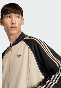 Beežikas võrgukangast lukuga jakk mustade detailidega, millel on õlgadel kolm musta triipu ja rinnal tikitud must logo.