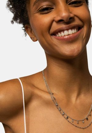 Zilveren gelaagde ketting met kleine ronde en ovale hangers. Draagt een witte spaghettibandtop tegen een effen achtergrond.