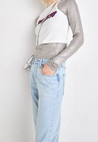 Person som bär en lager på lager silvrig långärmad topp under en vit ärmlös skjorta, ljusblå högmidjade jeans och bär en liten metallisk väska.