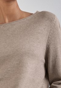 Närbild av en person som bär en beige stickad tröja med en bred, något avskuren halsringning mot en enfärgad bakgrund.