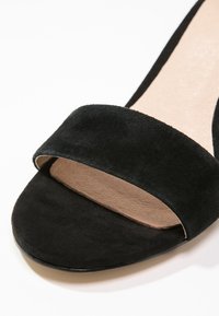 Gros plan d'une sandale en daim noir à bout ouvert avec une large bride avant et une semelle intérieure beige.