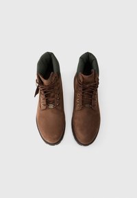 Botas de ante marrón con un collar acolchado verde, ojales metálicos y cordones marrones. Suelas de goma negra gruesa para mayor tracción.