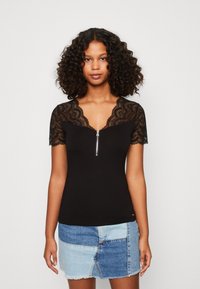 Schwarzes Oberteil mit kurzen Spitzenärmeln und Reißverschluss vorne. Strukturierte Spitze an den Schultern, enganliegende Silhouette, kombiniert mit einem Jeans-Patchwork-Rock.
