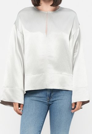 Vrouw draagt een witte satijnen blouse met lange mouwen, brede manchetten en een kleine voorste split, gecombineerd met blauwe denim jeans.