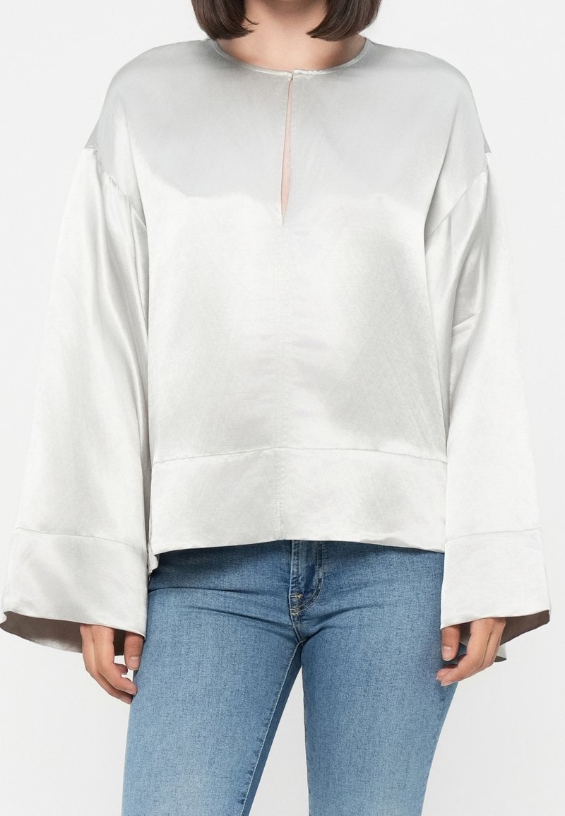 Femme portant un chemisier en satin blanc à manches longues avec de larges poignets et une petite fente sur le devant, associé à un jean en denim bleu.