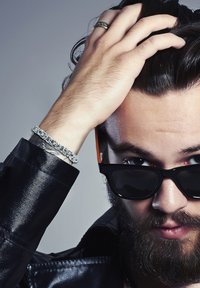 Sidabrinė grandinėlė ant riešo, juoda odinė striukė, tamsūs akiniai nuo saulės su oranžiniais akcentais, išbarstyti plaukai ir matoma barzda.