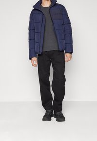 Marinblå pufferjacka med quiltat mönster, grå crewneck-tröja, svarta byxor och svarta skor med strukturerade sulor. Calvin Klein-logotyp på jackan.