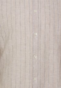 Světle béžová košile s vertikálními šedými pruhy, s knoflíkovým zapínáním vpředu a průhlednými knoflíky. Lněná textura s hladkým povrchem.