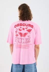 Rosa Übergröße T-Shirt mit roten Grafiken, die die Wörter "OBSCURE FUTURE" und Schmetterlingsillustrationen auf dem Rücken zeigen. Kurze Ärmel und lockere Passform.