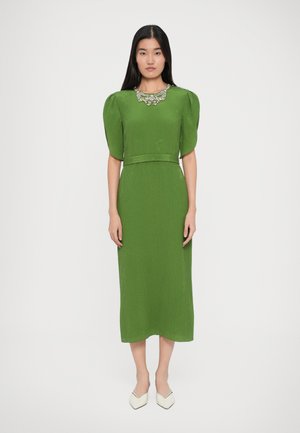 Robe midi verte à manches bouffantes et taille cintrée. Encolure ornée de détails floraux. Texture lisse, accompagnée de chaussures blanches à bouts pointus.