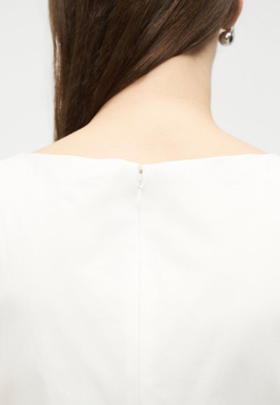 Γυναίκα με μακριά καστανά μαλλιά που φοράει ένα λευκό ένδυμα με φερμουάρ στην πλάτη και ένα ασημένιο σκουλαρίκι με καρφίτσα.