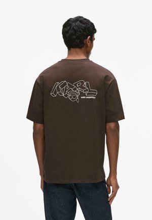 Muž v hnedom tričku s veľkým bielym nápisom „KARL“ a logom Karl Lagerfeld na chrbte, v kombinácii s tmavomodrými džínsami, otočený chrbtom.