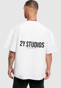 Baltas oversized marškinėliai trumpomis rankovėmis, su drąsiais juodais užrašais ant nugaros, kuriuose rašoma "2Y STUDIOS" ir papildomais mažais užrašais žemiau.