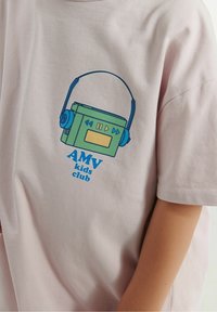 Šviesiai rožinė medvilninė marškinėliai su žalia boombox grafika, mėlynomis ausinėmis ir mėlynai užrašyta „AMV vaikų klubas“ apačioje.