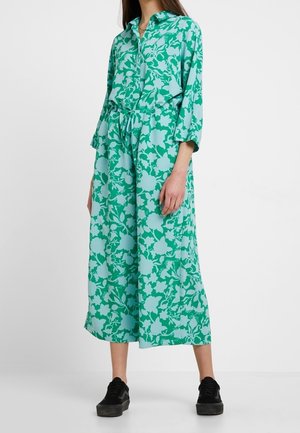 Femme portant une robe midi fleurie verte et bleu clair avec des manches trois-quarts, un col, une patte de boutonnage sur le devant et des baskets noires.
