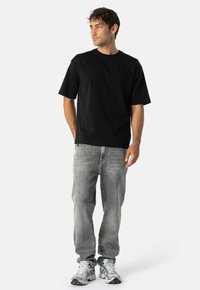 Melnas īsās piedurknes T-krekls no kokvilnas ar brīvu piegriezumu un apaļu izgriezumu, kombinēts ar gaiši pelēkiem džinsiem un pelēkiem sporta apaviem.