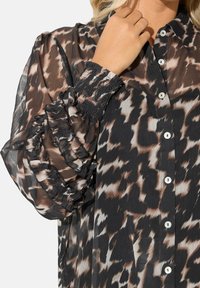 Camicia trasparente con bottoni presenta una stampa animale nera e marrone, maniche arricciate con volant e bottoni bianchi lungo la parte anteriore.
