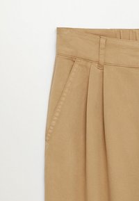 Pantalon beige taille haute avec plis devant, poches latérales et passants pour ceinture, présenté sur fond blanc.