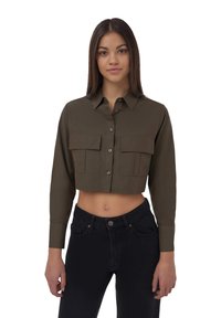 Camicia verde oliva a bottoni, tagliata, con due tasche sul petto e maniche lunghe; abbinata a jeans scuri a vita alta.