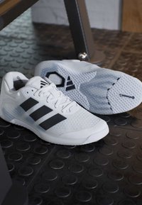 Λευκά αθλητικά παπούτσια με μαύρες ρίγες Adidas τοποθετημένα σε υφή μαύρου πατώματος γυμναστηρίου κάτω από εξοπλισμό άσκησης.