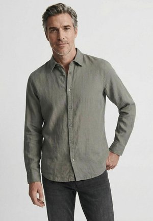 Mann mit grauen Haaren, der ein graues langärmeliges Hemd mit Knöpfen und schwarze Hose trägt, steht mit einer Hand auf der Hüfte vor einem schlichten Hintergrund.