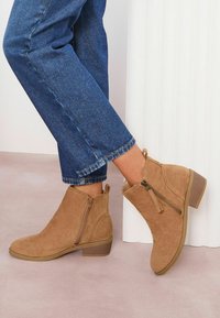 Botas de tobillo de ante en color beige con cremallera lateral, tacón bajo y apilado, y costuras decorativas; combinadas con jeans azules remangados.