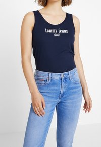 Marinblå linne med vit "tommy jeans" logo, parat med ljusblå denimjeans med dragkedja och klassisk 5-fickdesign.