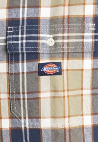 Poche de chemise à carreaux avec bouton blanc et étiquette de marque Dickies en rouge, bleu et jaune sur tissu à motif à carreaux orange, marine, beige et blanc.