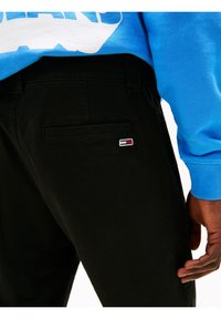 Černé bavlněné kalhoty s hladkou texturou, s zadní kapsou a malou nášivkou v červené, bílé a modré barvě. Modrá mikina částečně viditelná.