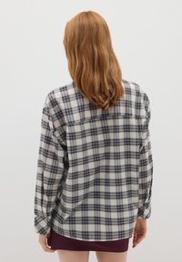 Persona con capelli rossi di lunghezza medio-lunga indossa una camicia larga a scacchi nera e bianca e una gonna corta di un viola scuro, con la schiena rivolta verso la macchina fotografica.