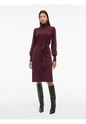 Bordo megztinis su aukštu kaklu, ilgomis pūstomis rankovėmis ir juosmeniniais diržais, derinamas su juodais aukštakulniais batais iki kelių.