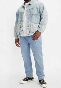 Man som bär en ljusblå denimjacka, vit t-shirt, ljusblå jeans och svarta canvas sneakers stående mot en enkel bakgrund.