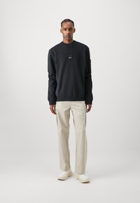 Felpa nera con collo arrotondato e piccolo patch con logo, abbinata a pantaloni beige chiaro. L'outfit è completato da sneakers bianche.