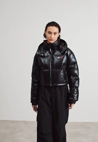 Chaqueta de plumas negra con cuello alto, textura brillante, cierre de cremallera y bolsillos frontales. Presenta un parche con el logo en el pecho. Se lleva con pantalones negros.