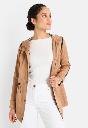 Femme portant une veste beige avec une doublure de capuche en imprimé léopard, une chemise blanche et un pantalon blanc, regardant vers sa droite contre un fond uni.