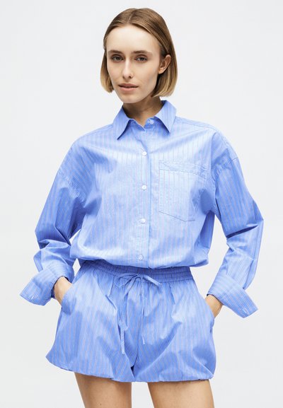 Νεαρή γυναίκα με κοντά μαλλιά που φοράει ανοιχτό μπλε ριγέ μακρυμάνικη μπλούζα και σορτς με κορδόνι ταιριαστά, με τα χέρια στις τσέπες, ουδέτερο φόντο.