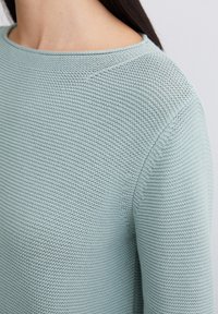 Světle modrý pletený svetr s texturovaným vzorem, širokým kulatým výstřihem a uvolněným střihem. Materiál vypadá měkký a prodyšný.