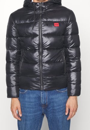 Schwarze Steppjacke mit glänzendem Finish, durchgehendem Reißverschluss, Kordelzugkapuze und einem kleinen roten Logo-Patch oben links. Strukturierte, gesteppte Optik.