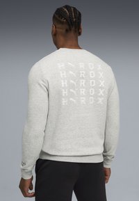 Sweat à capuche gris avec un dos à motifs comportant le texte répété "HYROX" et un logo. Poignets et ourlet côtelés, associé à un pantalon noir.