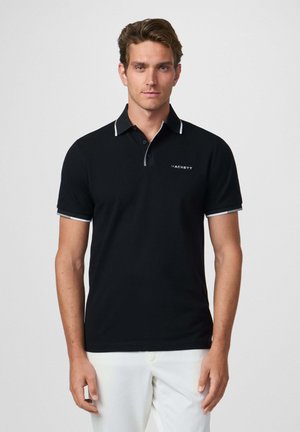 Camisa polo negra con acentos blancos, de manga corta y cuello de dos botones, con un logo en el lado izquierdo del pecho. Textura suave.