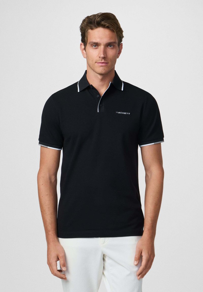 Camisa polo negra con acentos blancos, de manga corta y cuello de dos botones, con un logo en el lado izquierdo del pecho. Textura suave.