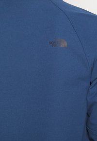 Tessuto blu testurizzato con il logo nero di The North Face sulla parte superiore della schiena, che mostra i dettagli delle spalle e una parte della cucitura del braccio.