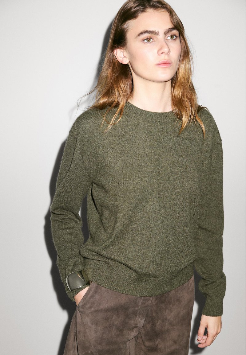 Maglione verde oliva con scollo rotondo e maniche lunghe, realizzato in un morbido materiale a maglia, abbinato a pantaloni in suede marroni.