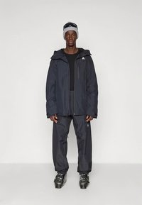 Άνδρας που φορά μαύρο μπουφάν σκι, παντελόνια, γκρίζο σκουφί με γυαλιά ηλίου από πάνω, και μαύρες μπότες σκι, στέκεται σε ουδέτερο φόντο.