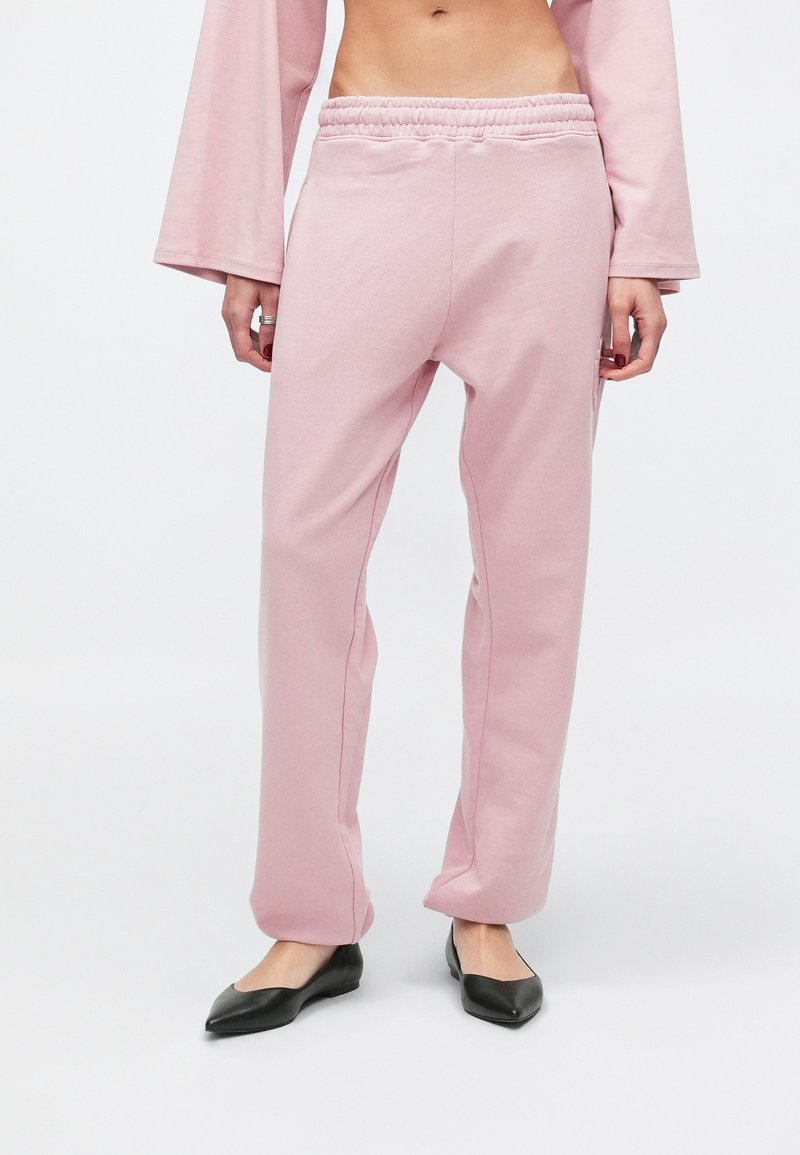 Calças de fato de treino cor-de-rosa com ajuste descontraído, cintura elástica e detalhes nas costuras laterais, combinadas com sapatilhas pretas. Textura suave, estilo casual.